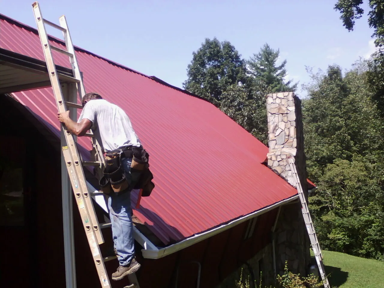 Roofer on a ladder beside a red metal roof and stone chimney — Soffit & Fascia Repair by Roofing Brothers in Balaton